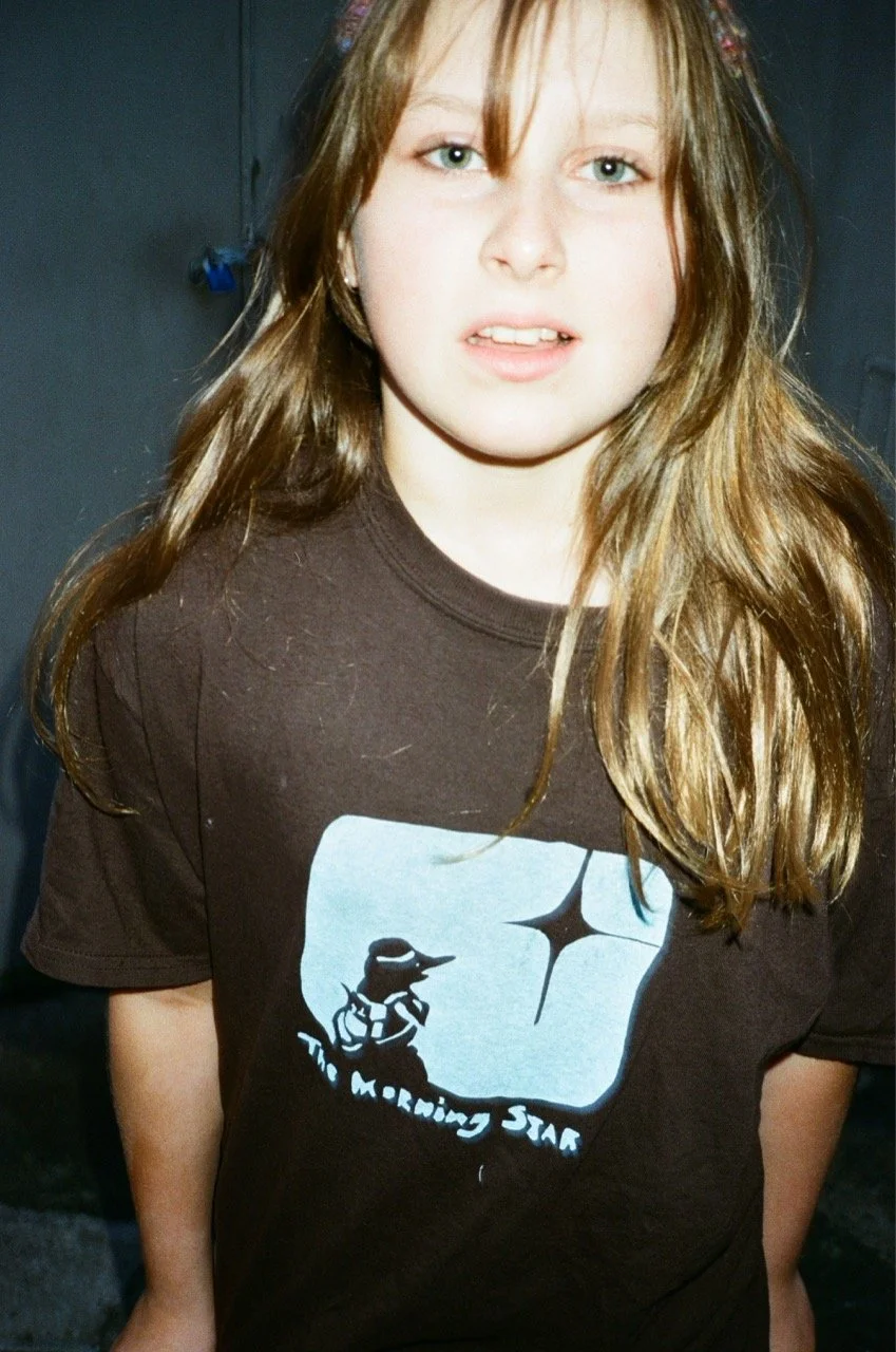 A young girl with long light brown hair, blue eyes, and fair skin wearing a black T-shirt with a graphic and text. The background is dark and indistinct.