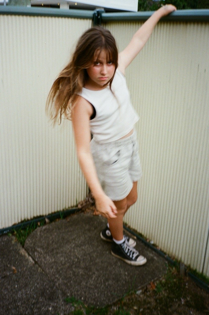 A girl with long brown hair, wearing a sleeveless white shirt, beige shorts, and black Converse sneakers, leans against a beige fence with an annoyed or serious expression.