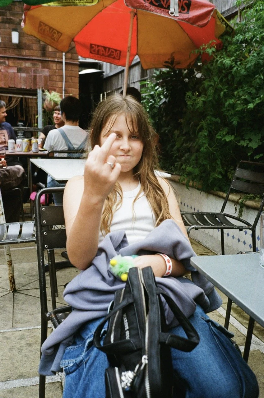 A girl sitting at an outdoor cafe giving a middle finger gesture towards the camera under a large red and yellow umbrella, with other people in the background.