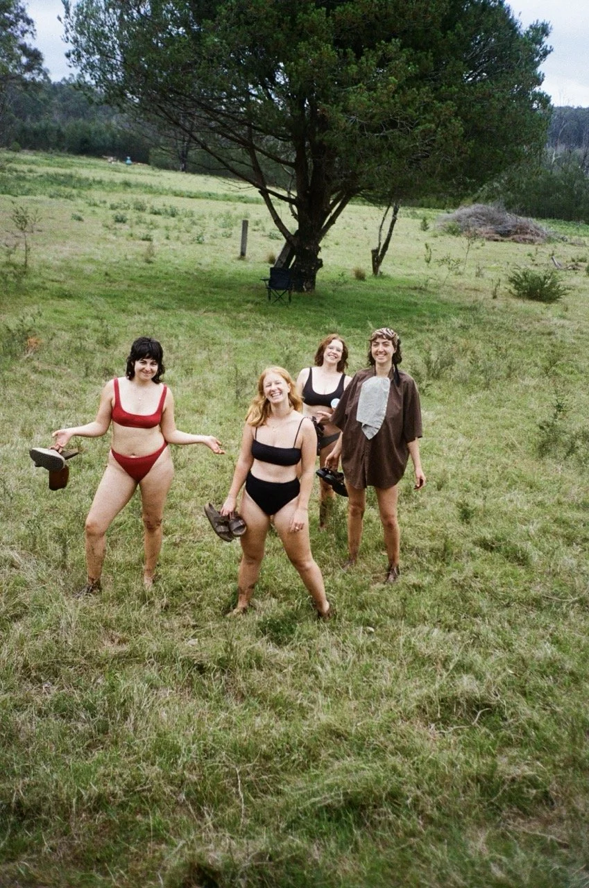 Four women standing in a grassy field, wearing minimal clothing, with trees and hills in the background. One woman is holding sandals, and another is wearing a brown shirt and gray tie. They are smiling and appear to be enjoying a casual outdoor mome