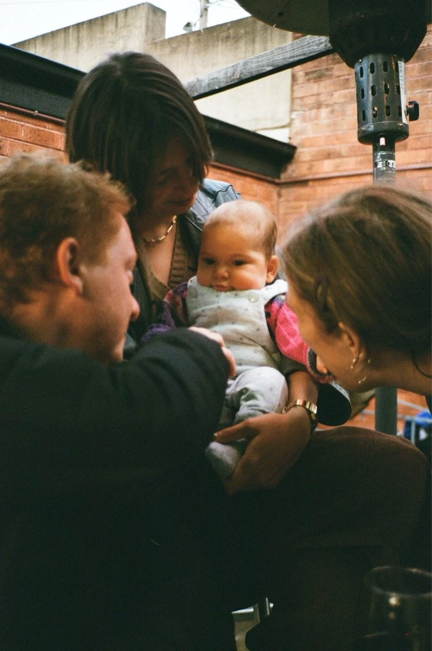 Four people, including an infant, are gathered closely together, seemingly engaged in a joyful moment, with the infant being held in the center. The setting appears to be outdoors, with brick walls and some wooden and metal structures visible.