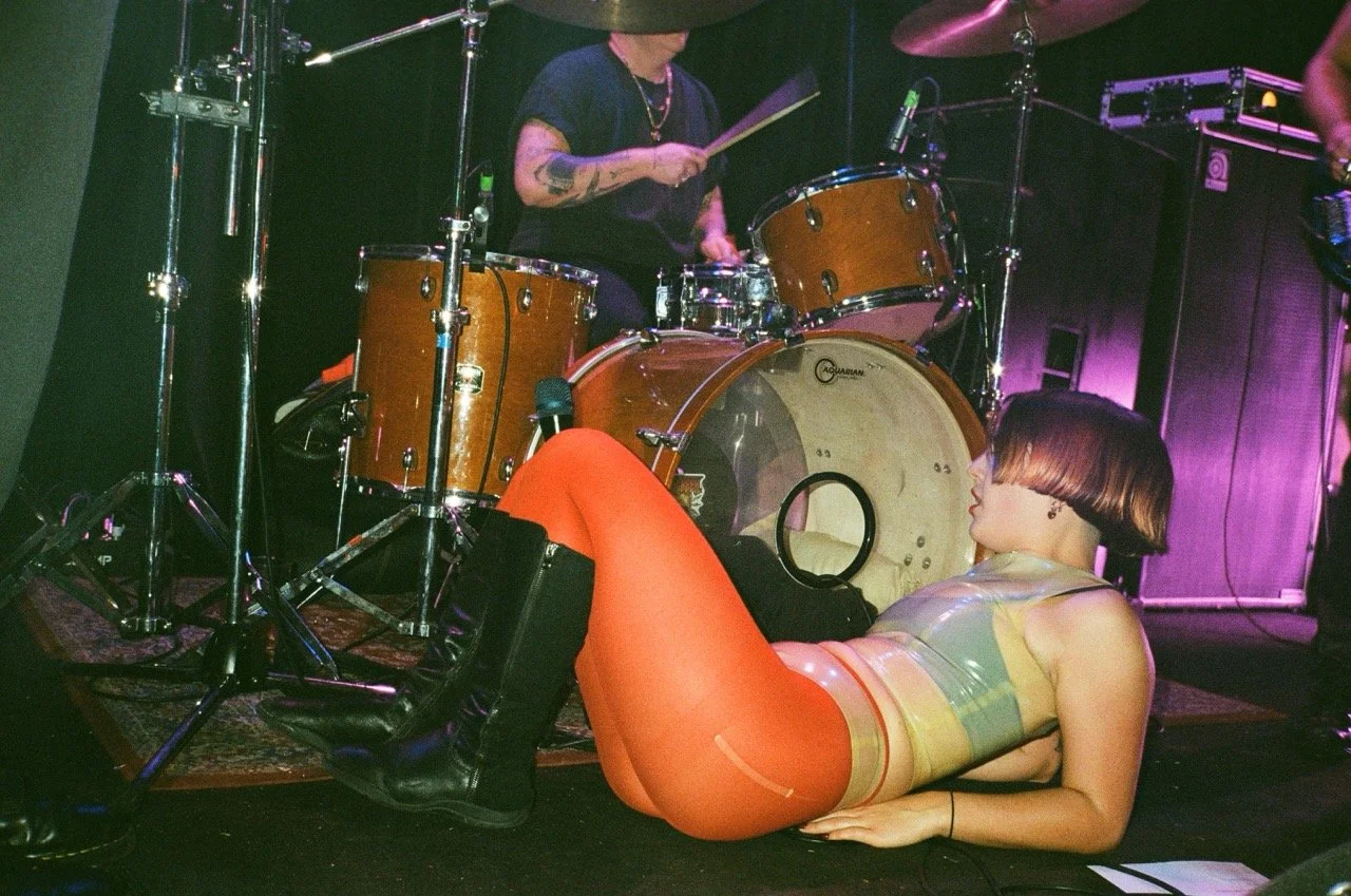 A woman with short hair, wearing orange pants and a translucent top, is lying on the floor near a drum set while a person behind the drums is playing. The scene appears to be in a music venue or stage setting with purple lighting.