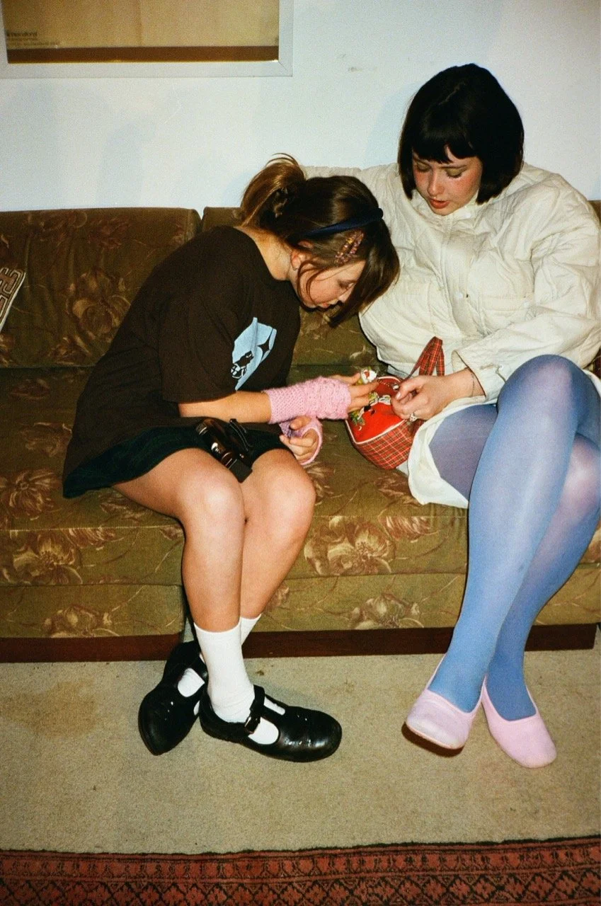 Two teenage girls sitting on a floral patterned couch, examining a small item together. The girl on the left has a ponytail, wearing a black T-shirt, white socks, and black shoes. The girl on the right has short black hair, wearing a white jacket, bl