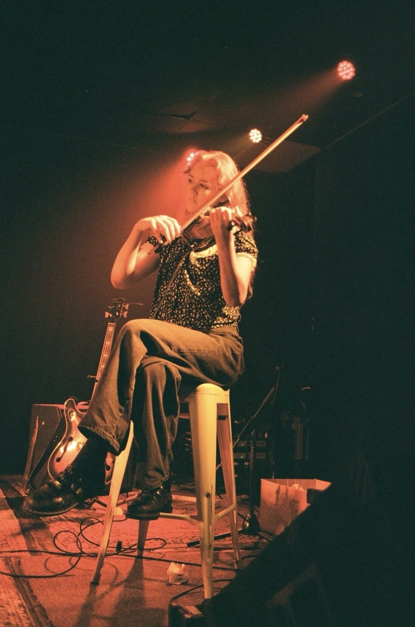 A woman playing the violin on stage, sitting on a high stool, with stage lights shining down on her.