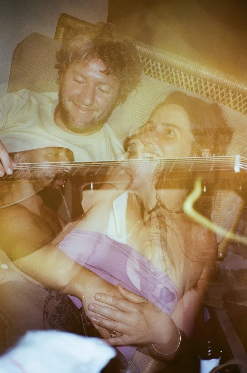 A man and woman enjoying each other's company, with the woman playing an acoustic guitar. The man is smiling and the woman is singing or talking, both sitting on a wicker outdoor furniture piece.