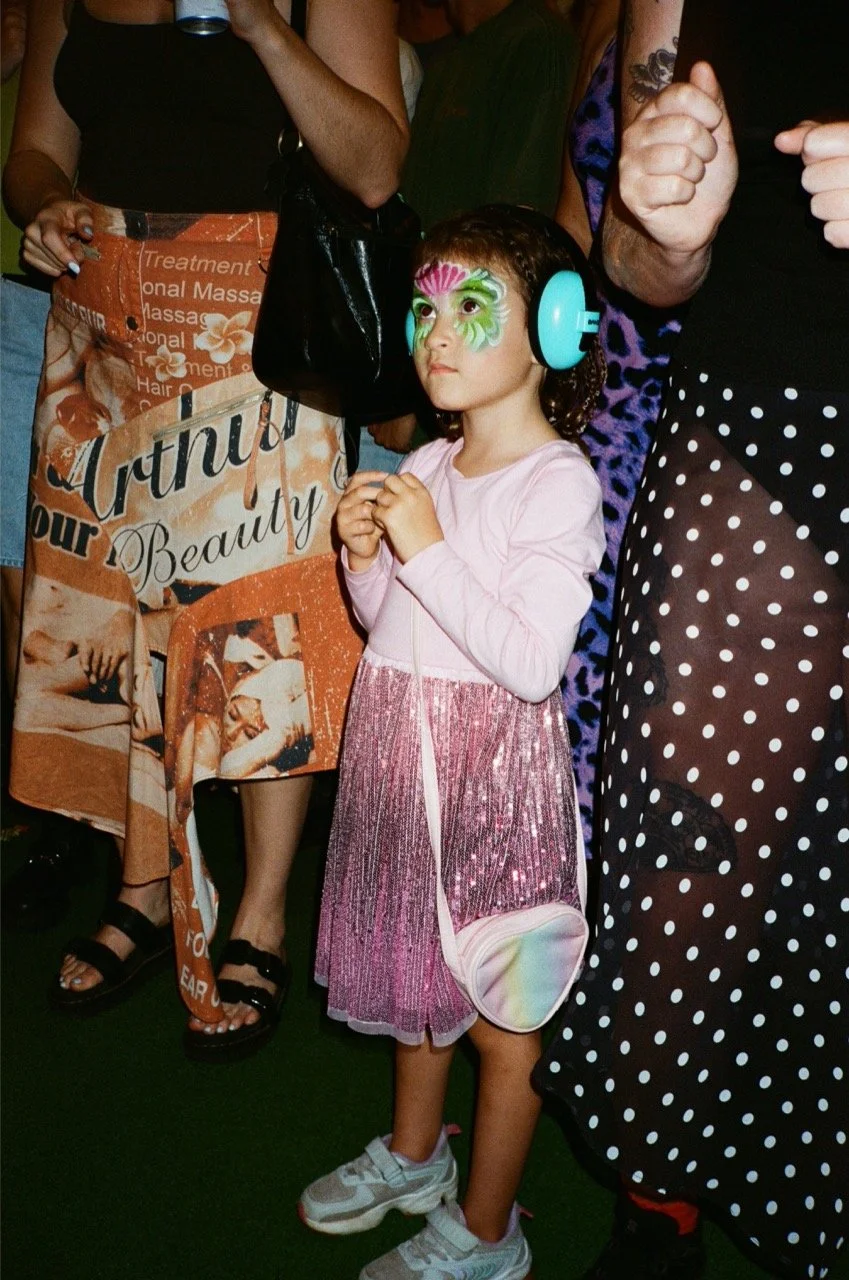 Young girl with floral face paint, wearing large teal headphones, pink and glittery dress, and sneakers, standing among adults at a social event.