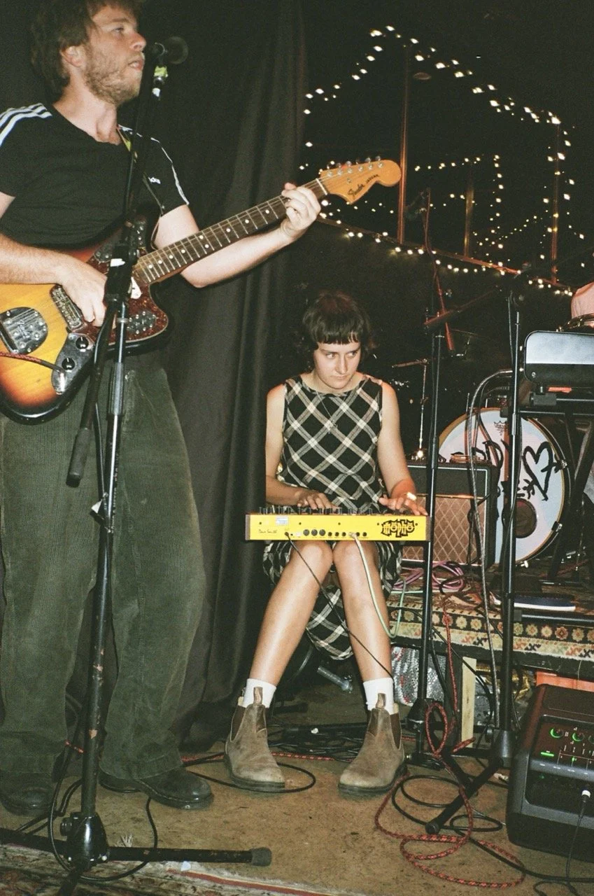 A musician playing an electric guitar on stage with a woman sitting and playing a yellow keyboard, with stage lights and musical equipment in the background.