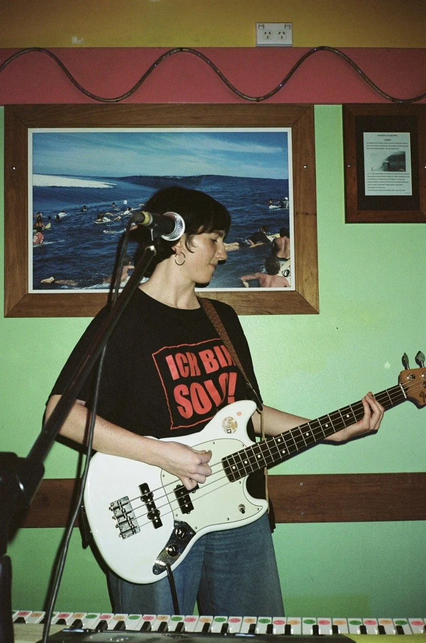 A young woman with short dark hair is playing a white electric bass guitar in a room with green walls. She is wearing a black t-shirt with red text that reads 'ICH BIN SOLZ!' and is standing in front of a framed ocean scene photograph. A microphone o