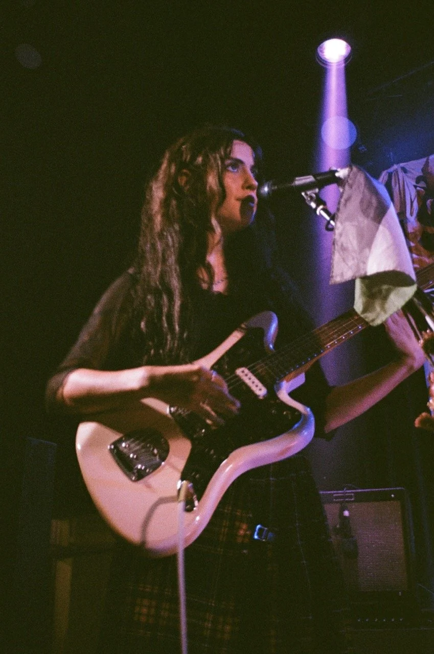 A woman with long wavy hair playing an electric guitar on stage under purple stage lights.