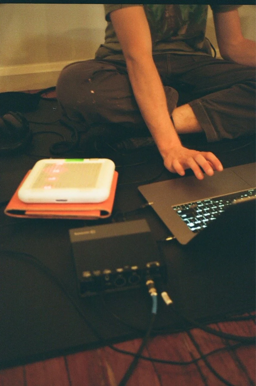 Person sitting cross-legged on the floor with a laptop, a smartphone on a case, and audio equipment on a black mat.