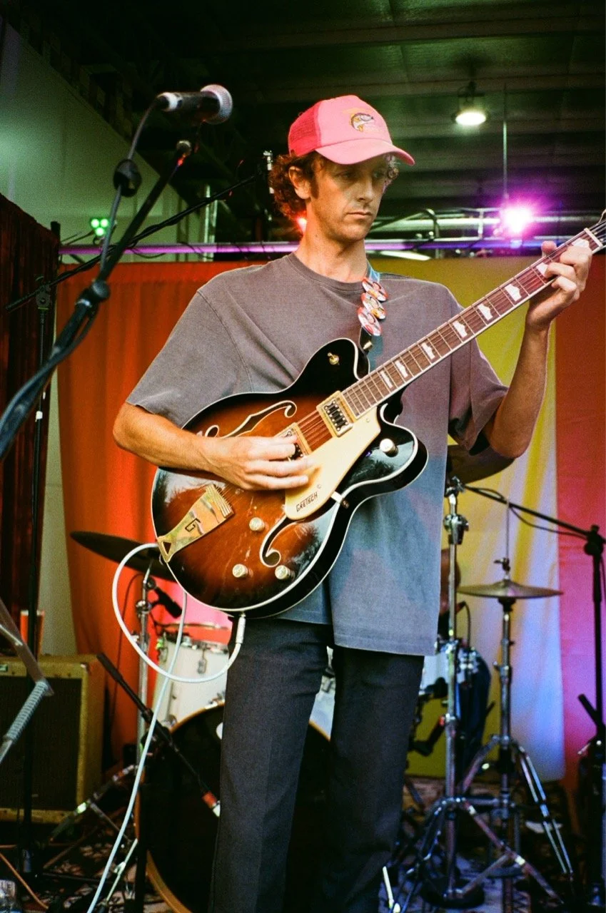 Man in a pink hat playing a hollow body electric guitar on stage with drum set and microphone nearby.