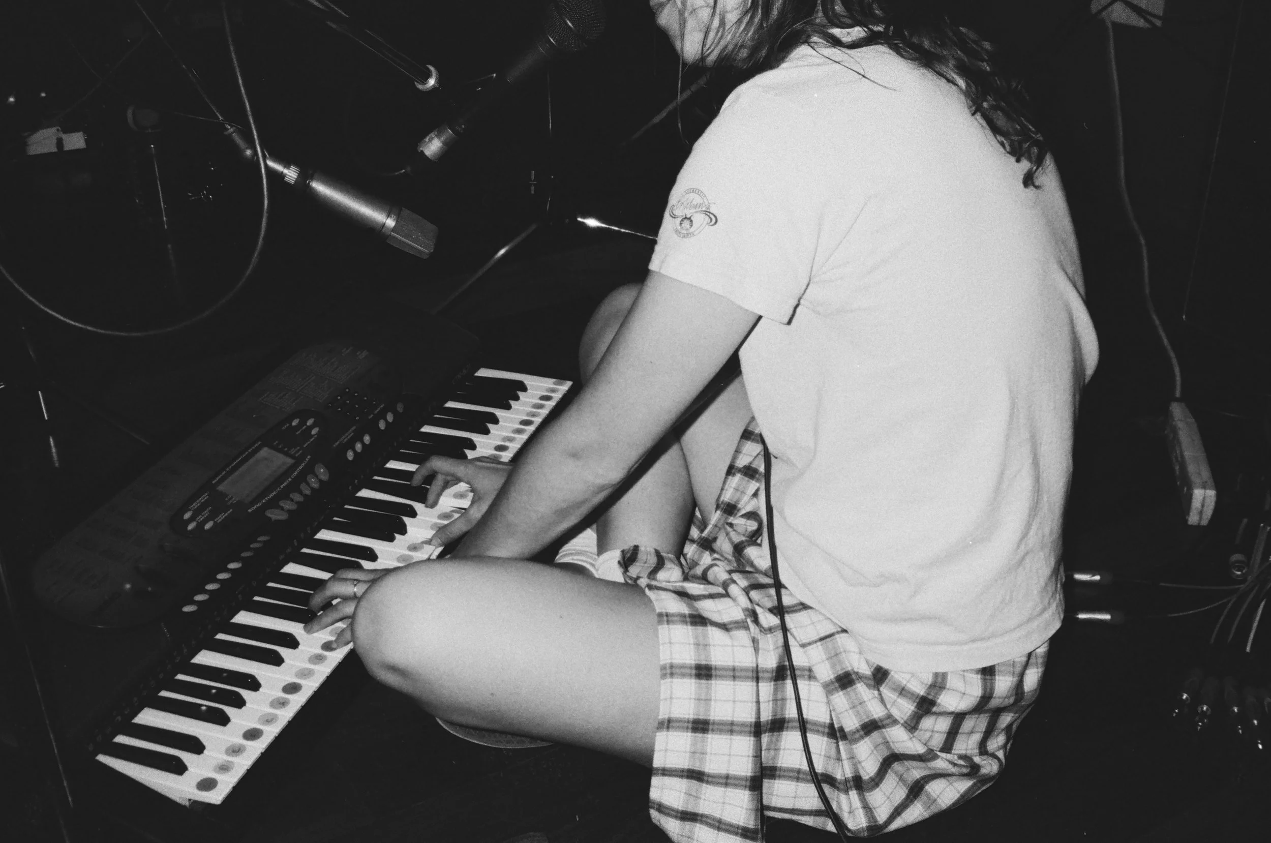 A person sitting cross-legged on the floor, playing a keyboard in a recording studio, surrounded by microphones and wires, wearing a plain light-colored t-shirt and checkered shorts.
