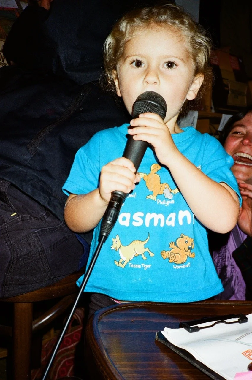 A young child with curly blonde hair holding a microphone, wearing a blue T-shirt with cartoon animals and the word "Tasman" on it, standing at a table.