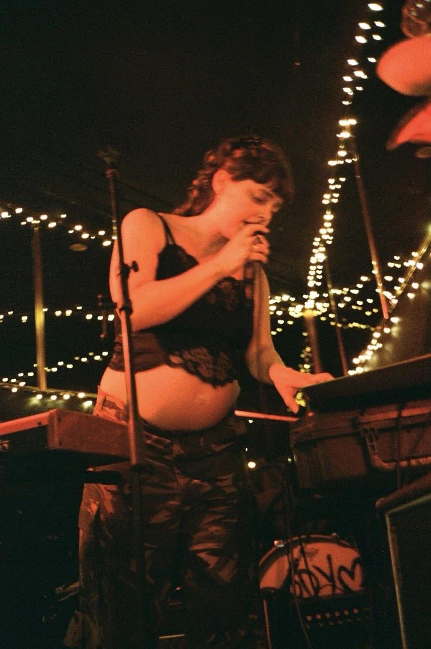 A pregnant woman singing into a microphone on stage at a dimly lit venue with string lights overhead.
