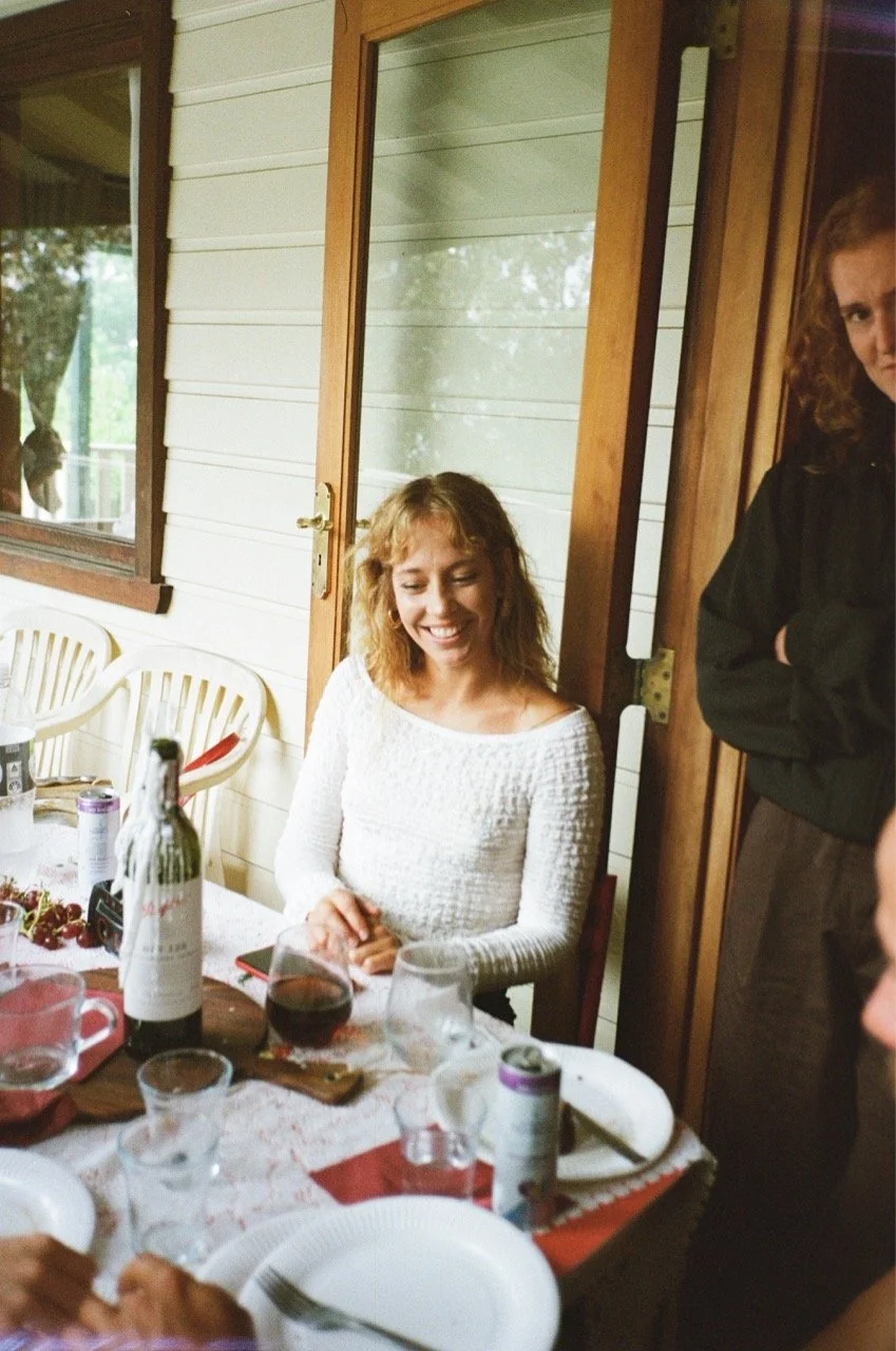 A woman with curly red hair smiling and sitting at a table set for a meal, with wine and other drinks, in a cozy outdoor patio area.