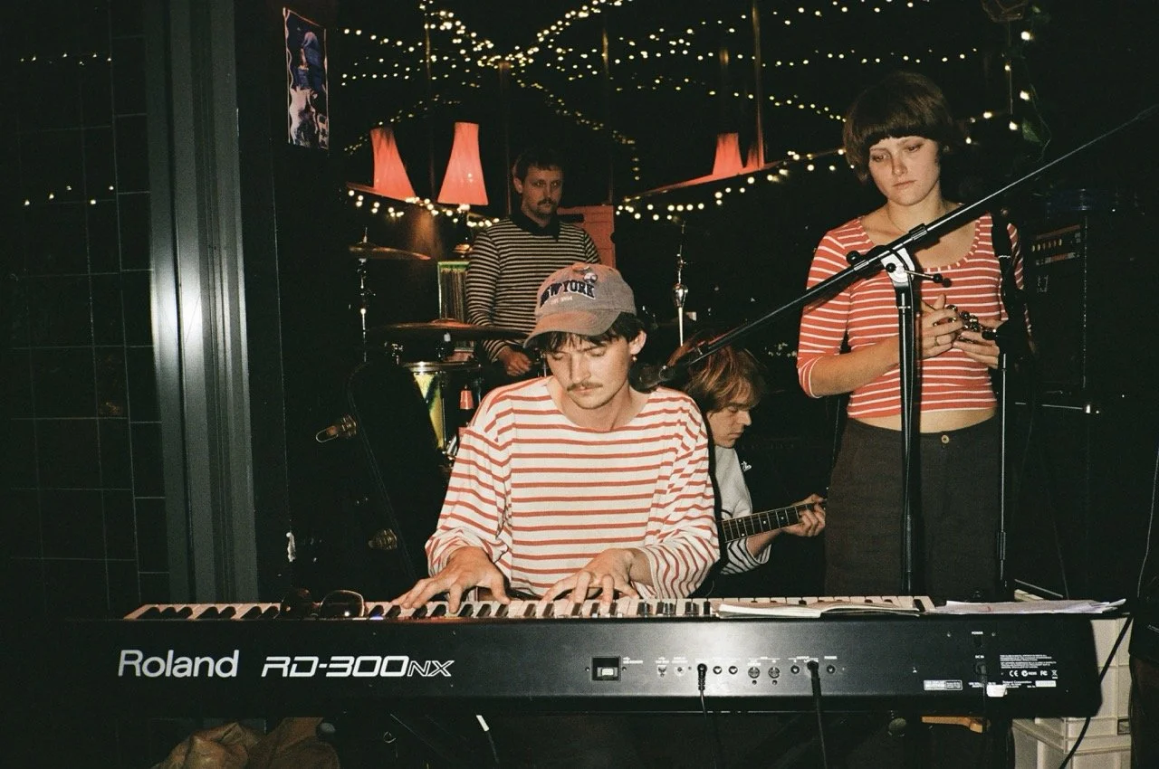 Four musicians performing with a keyboard, guitar, drums, and a vocalist, in a dimly lit room decorated with string lights and red lamps.