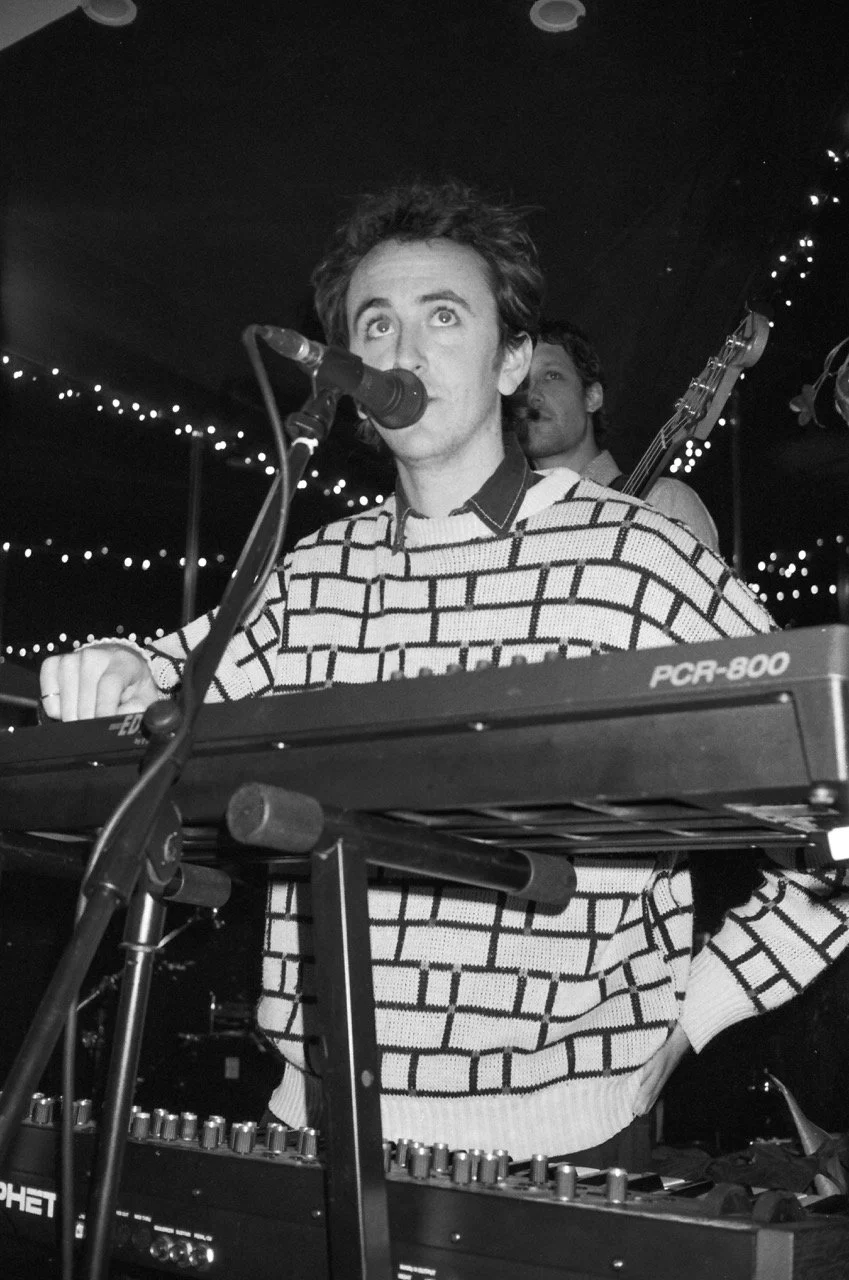 Black and white photo of a man singing into a microphone while playing an electronic keyboard, with string lights in the background.