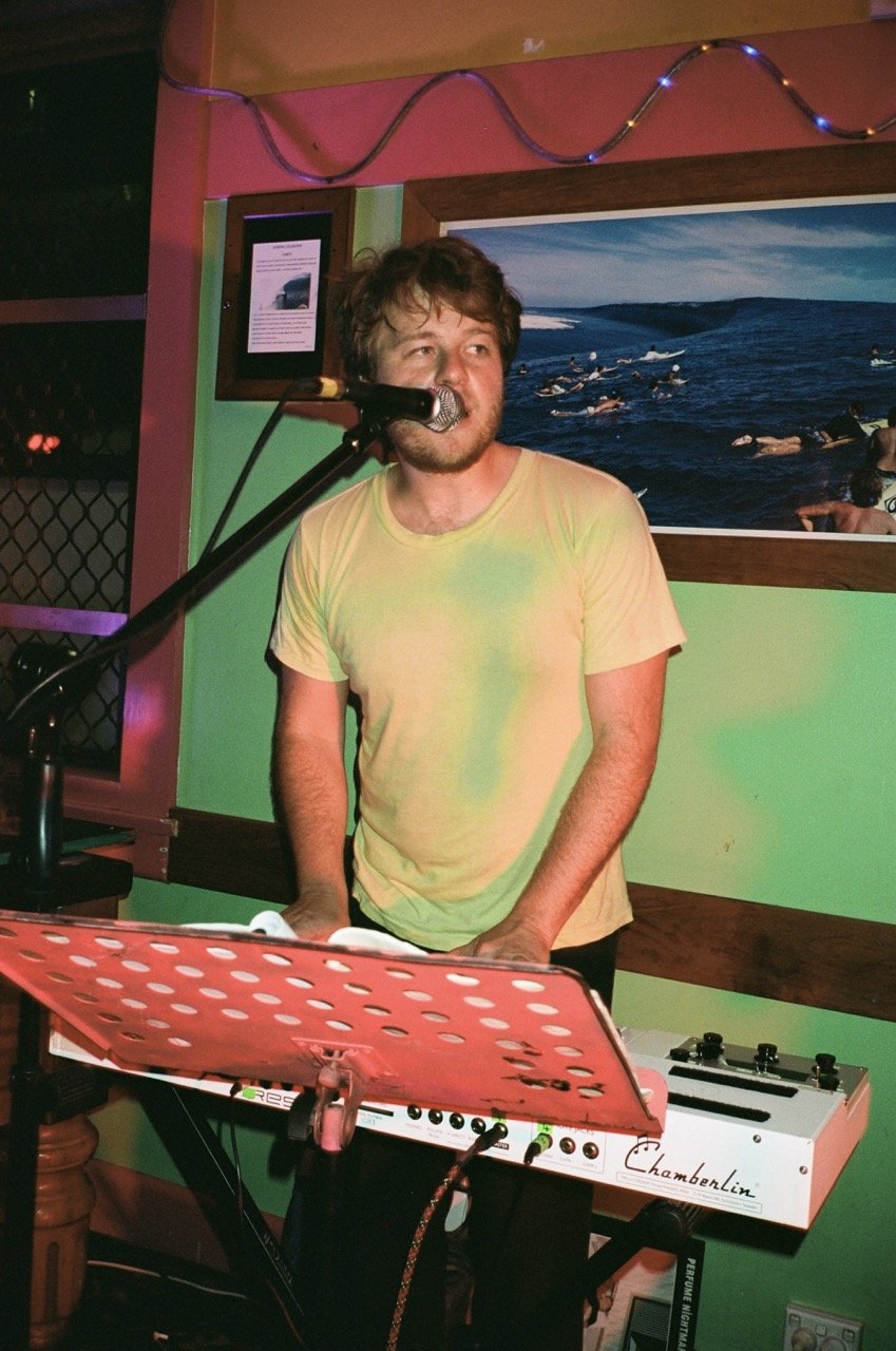 Man with brown hair and beard singing into a microphone while playing a keyboard in a colorful room with pink and green walls, a framed picture, and string lights