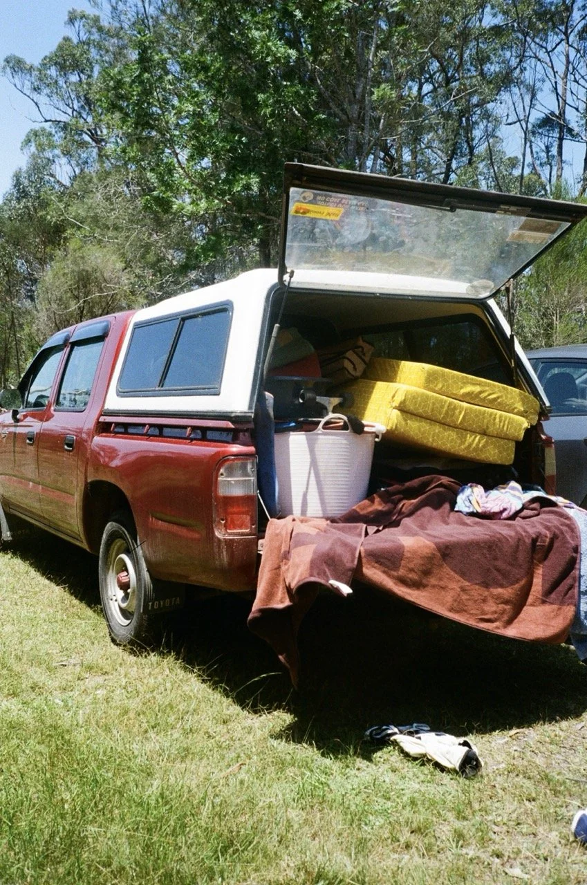 Red pickup truck with a white camper shell parked on grass, with its tailgate down, carrying camping gear including foam mattresses, a cooler, blankets, and clothing, surrounded by trees on a sunny day.
