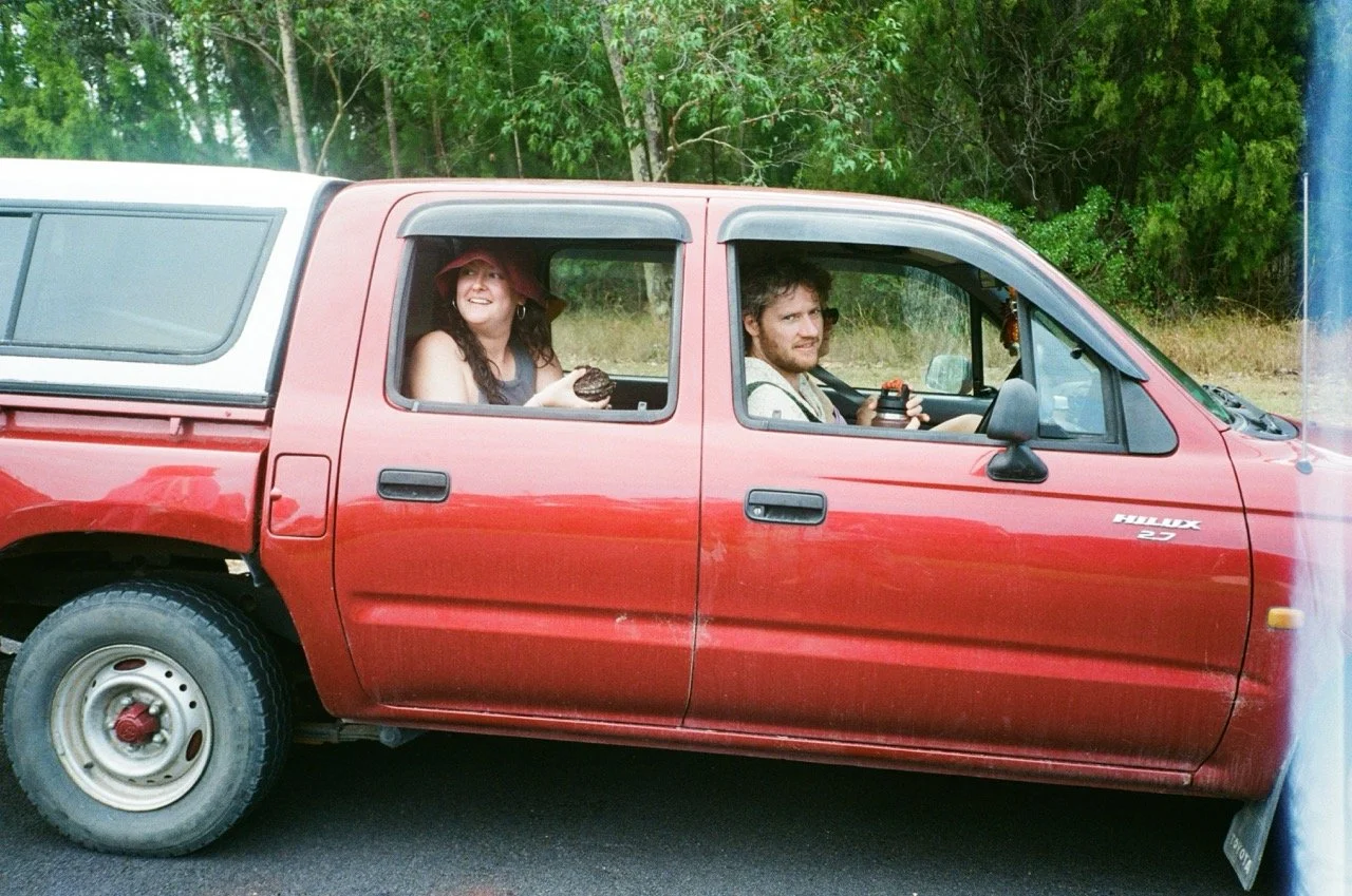 A red pickup truck with two people inside, a woman in the back seat holding a donut and a man in the front seat holding a coffee mug, parked on the side of a road near trees.