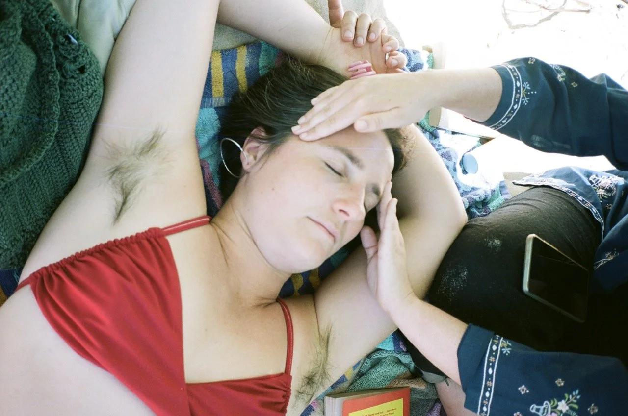 A woman with dark hair and tattooed armpit lying on her back with eyes closed, receiving a forehead and cheek massage from a person with crossed arms wearing a blue shirt with snowflake patterns.