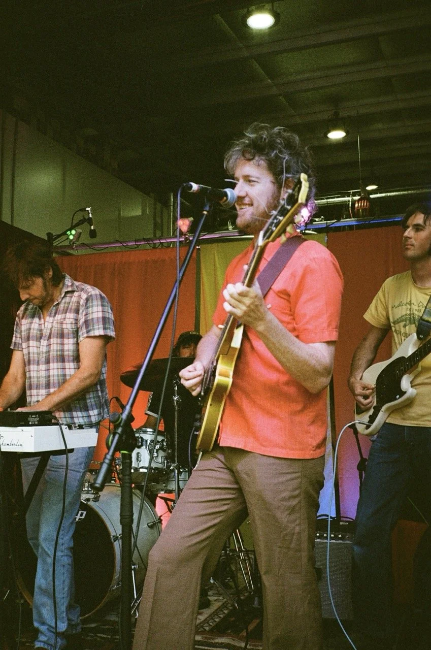 A band performing live on stage, featuring a man singing into a microphone and playing an electric guitar, with two other band members playing a keyboard and bass guitar, against an orange and yellow backdrop.
