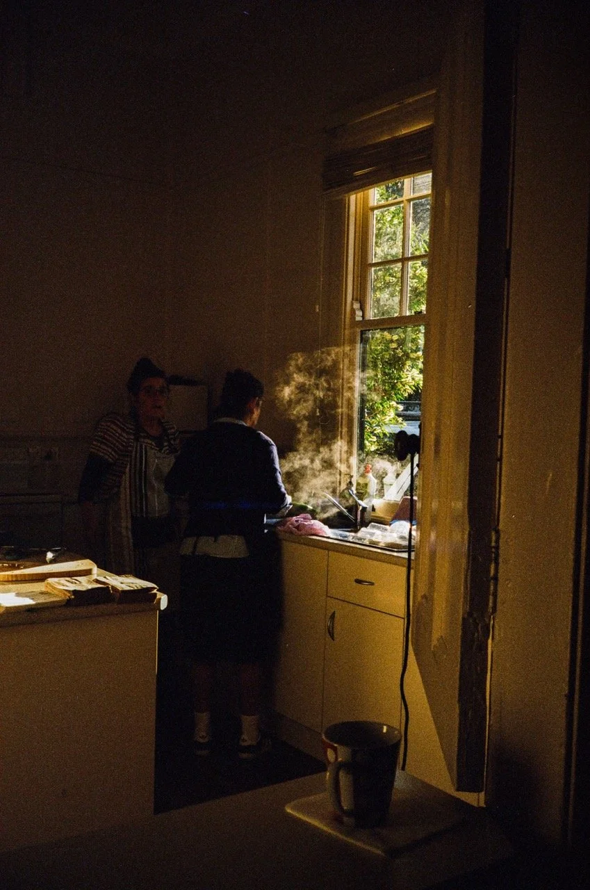 Two women in a kitchen near a window, with steam rising from a stove as sunlight streams in.