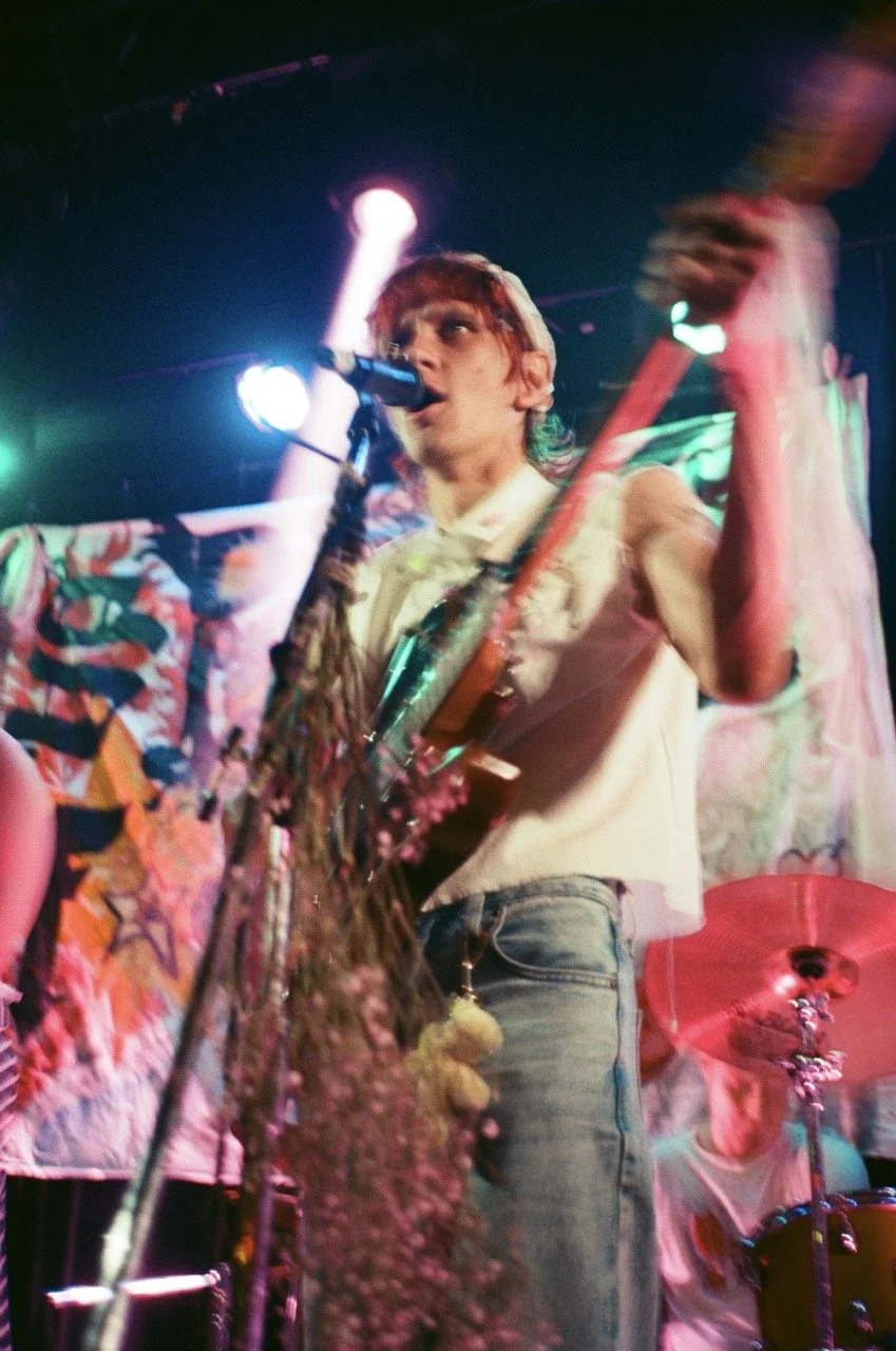 A musician with reddish hair playing an electric guitar and singing into a microphone on stage with colorful lighting, a person in the background playing drums, and a pink umbrella visible.
