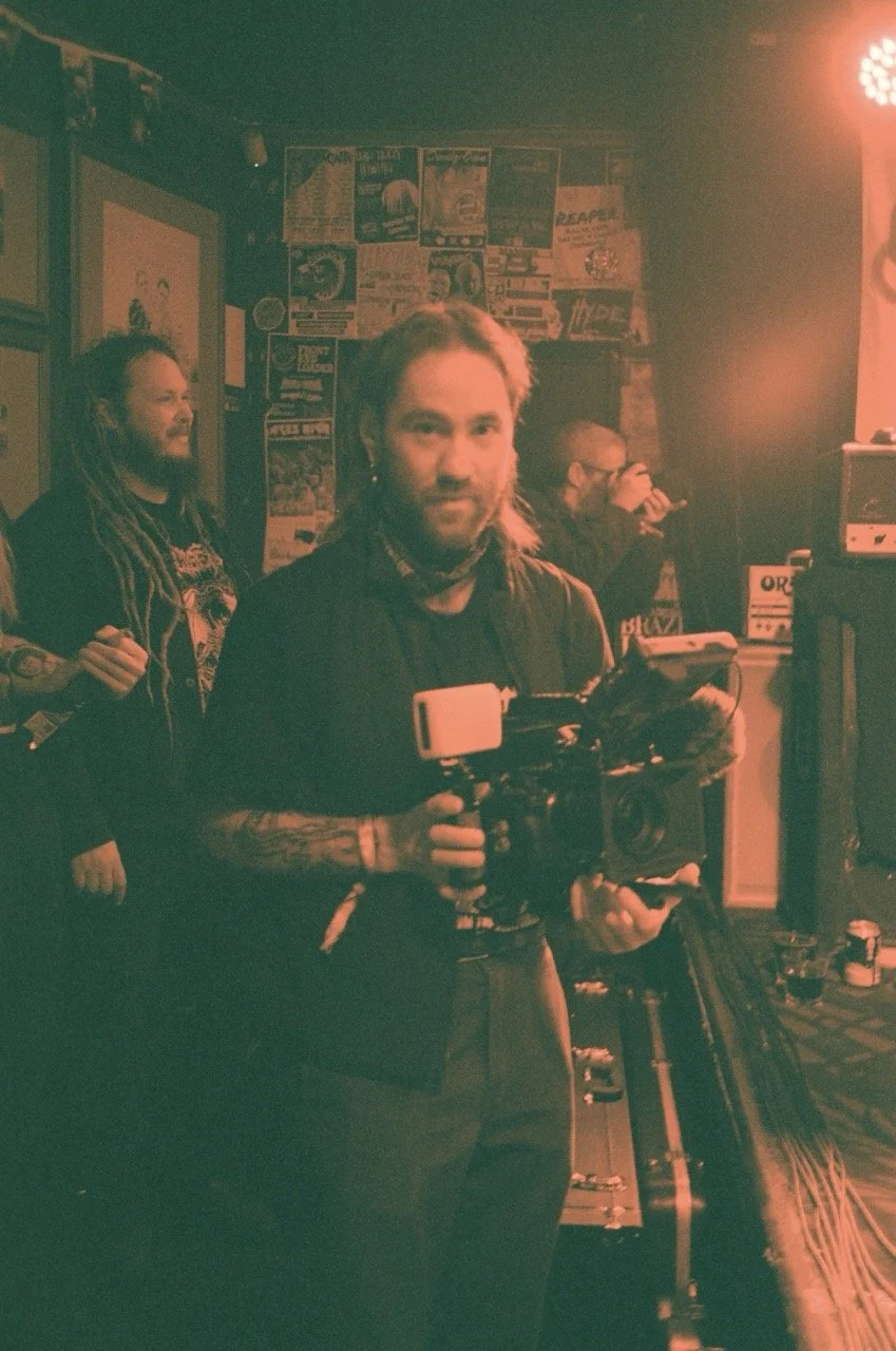 Man holding a video camera in a dimly lit room with posters on the wall and other people in the background.