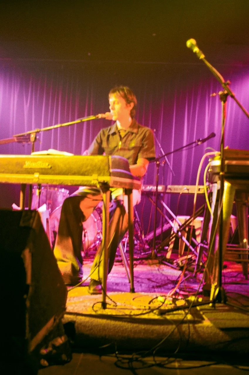 A young musician playing a keyboard on stage with purple curtains behind and multiple microphones and cables around.