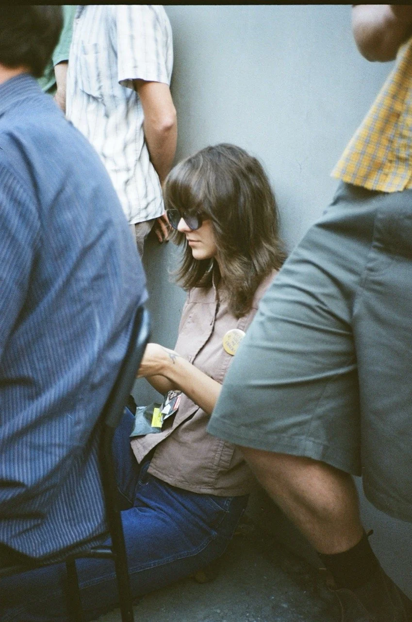 A woman with sunglasses and brown hair sitting on the floor, surrounded by several people standing around her.