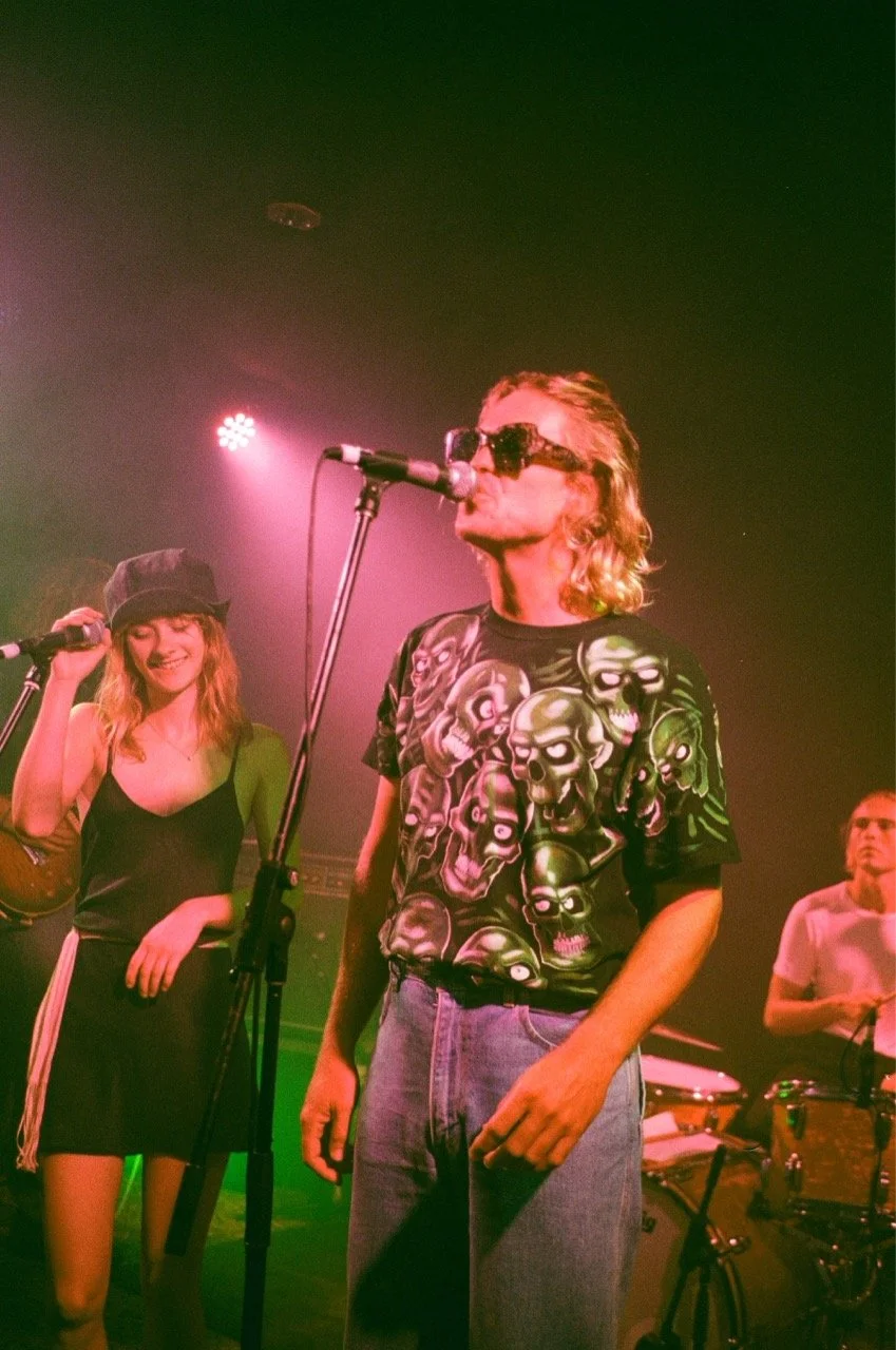 Band members performing on stage with colorful lighting, including a woman wearing a black dress and hat, and a man with long hair wearing sunglasses and a graphic skull shirt.