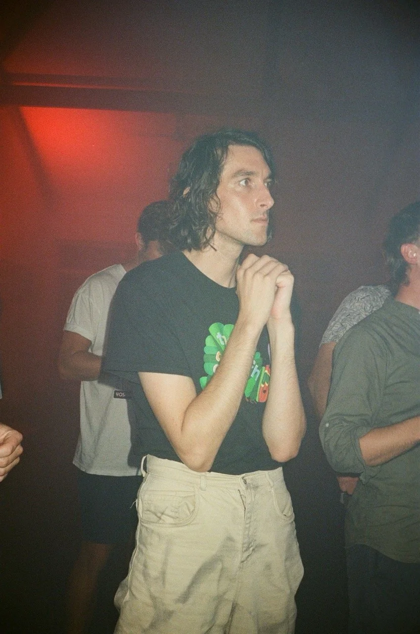 A young man with long, curly dark hair, wearing a black T-shirt with colorful graphics and beige pants, stands with hands clasped near his face, in a dimly lit room with a red light.