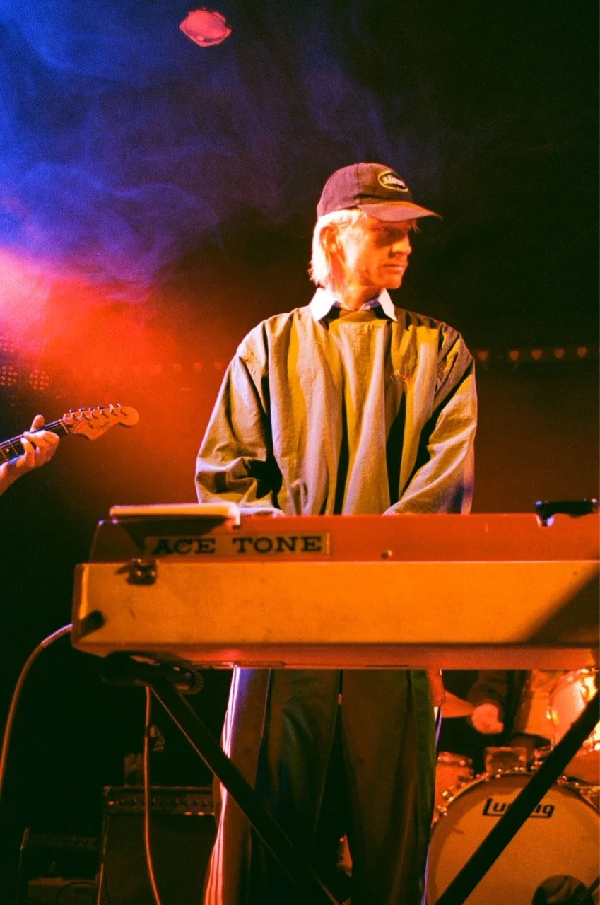 A musician with blonde hair wearing a baseball cap and oversized jacket playing a keyboard labeled 'ACE TONE' on stage, with colorful stage lighting and a drum set in the background.