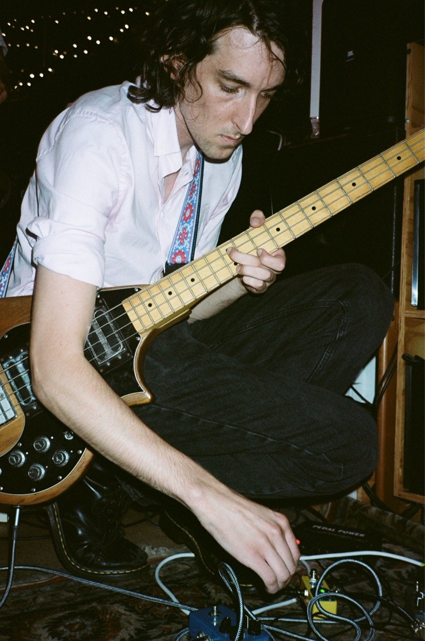 A young man with long curly hair is sitting on the floor, playing an electric guitar. He is wearing a white shirt with rolled-up sleeves and black pants, focusing intently on his guitar. There are various audio equipment and cables around him, with a