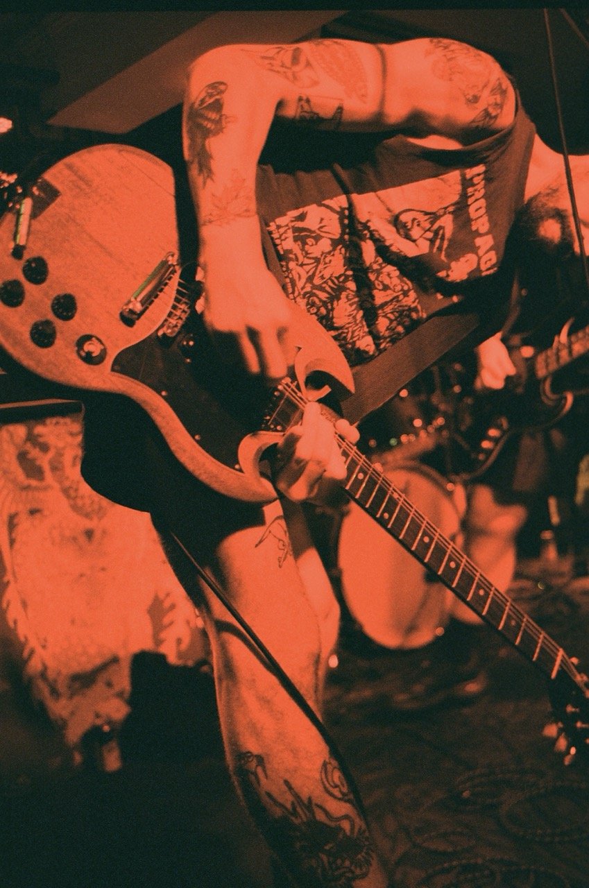 A person playing an electric guitar on stage, with tattoos visible on their arm and leg, under red stage lighting.