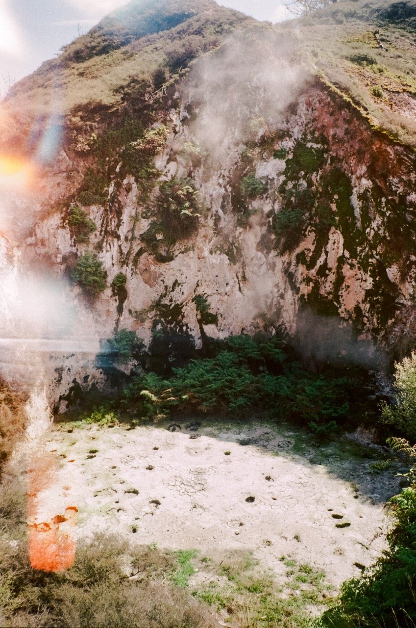 A dry, cracked, and arid riverbed surrounded by rocky cliffs and green vegetation at the base of a hillside.