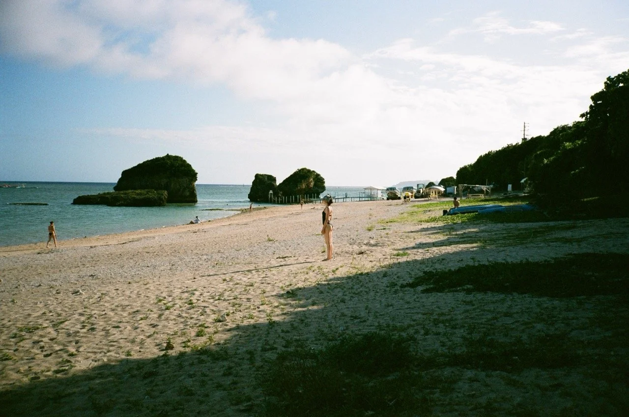 Beach scene with a woman standing on sand, ocean waves, large rocks in water, and some people in the distance. There are trees on the right side and a partly cloudy sky.