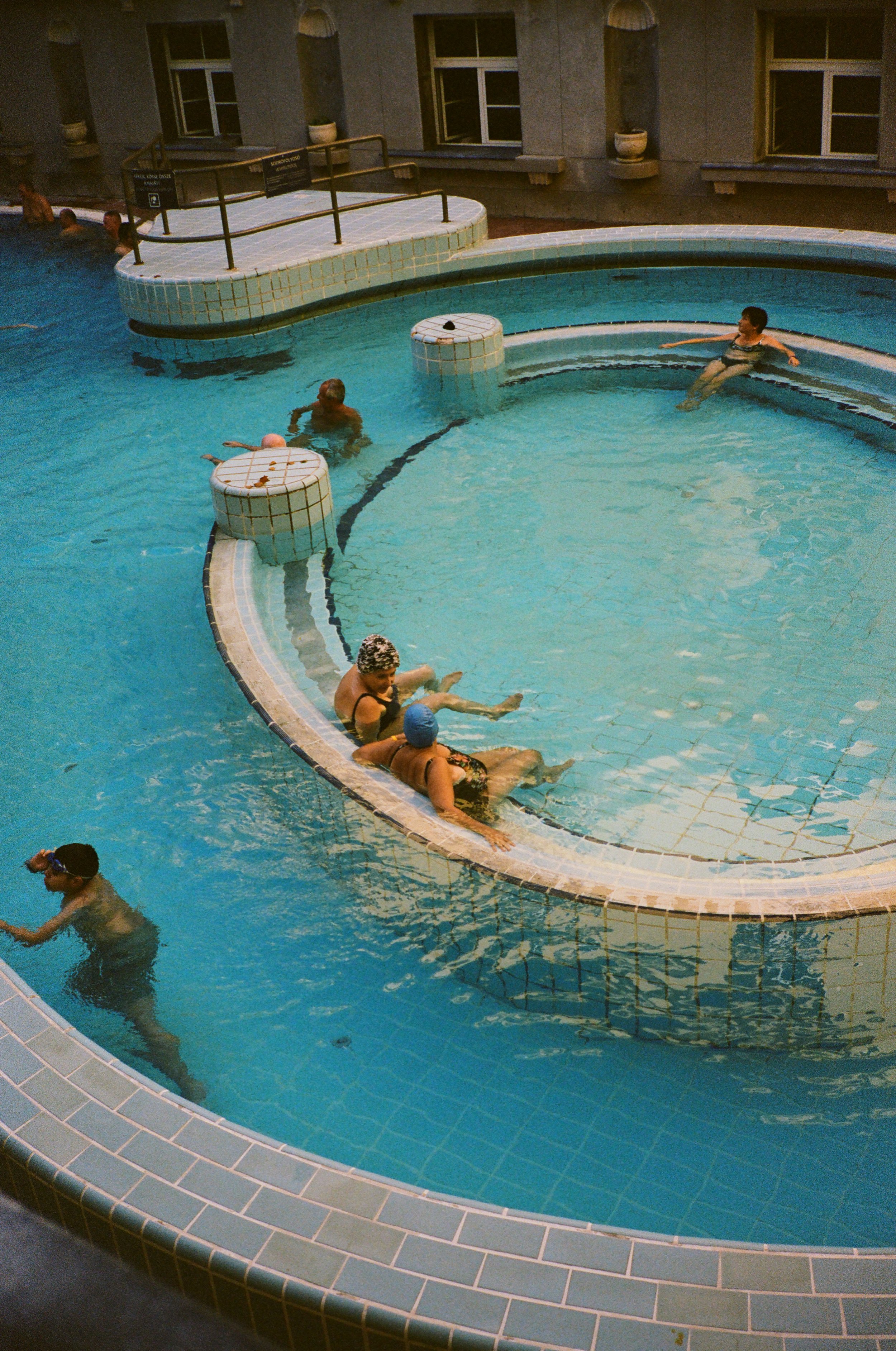 People relaxing and swimming in an indoor swimming pool, some sitting on the edge and others in the water.