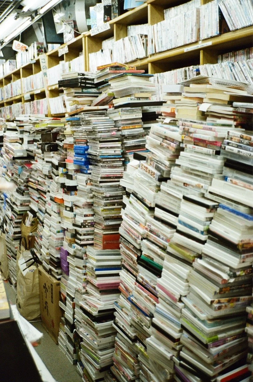 Stacks of scattered books and magazines in a cluttered bookstore aisle with shelves full of books in the background.