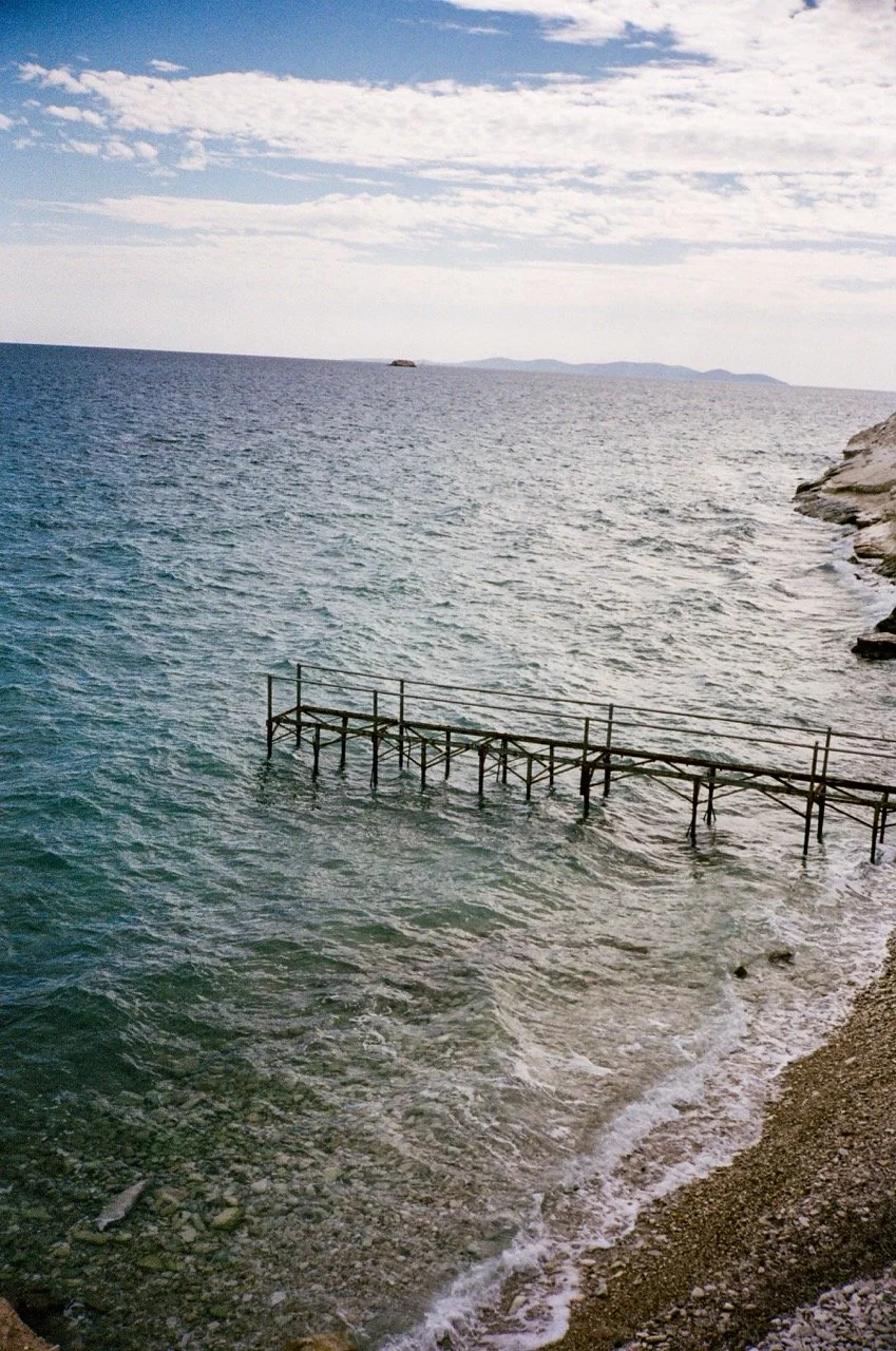 A wooden dock extending into the ocean with waves gently crashing on the rocky shore, under a partly cloudy sky.