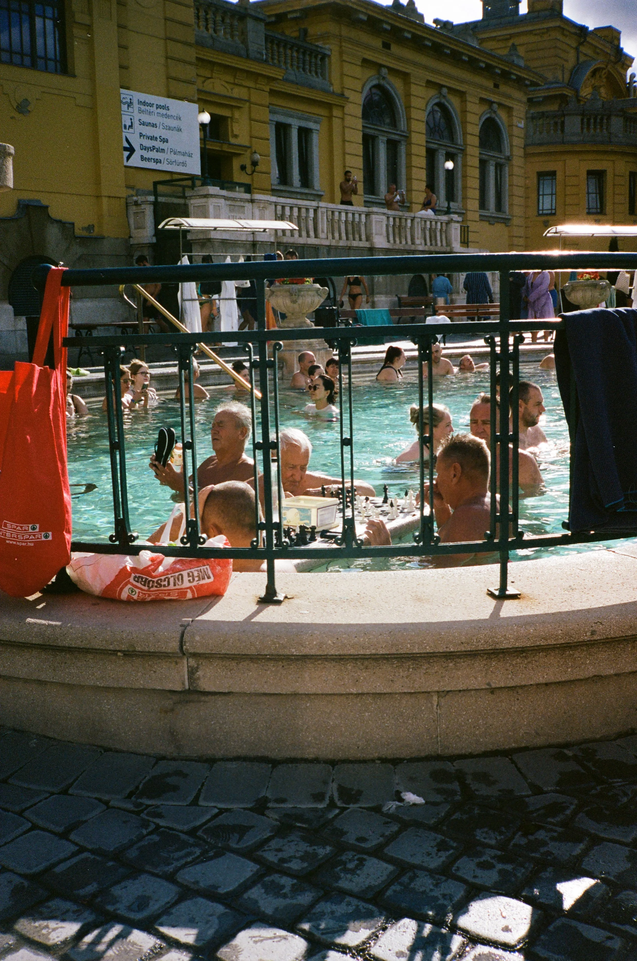 People swimming and relaxing in a public outdoor swimming pool with a decorative building in the background.