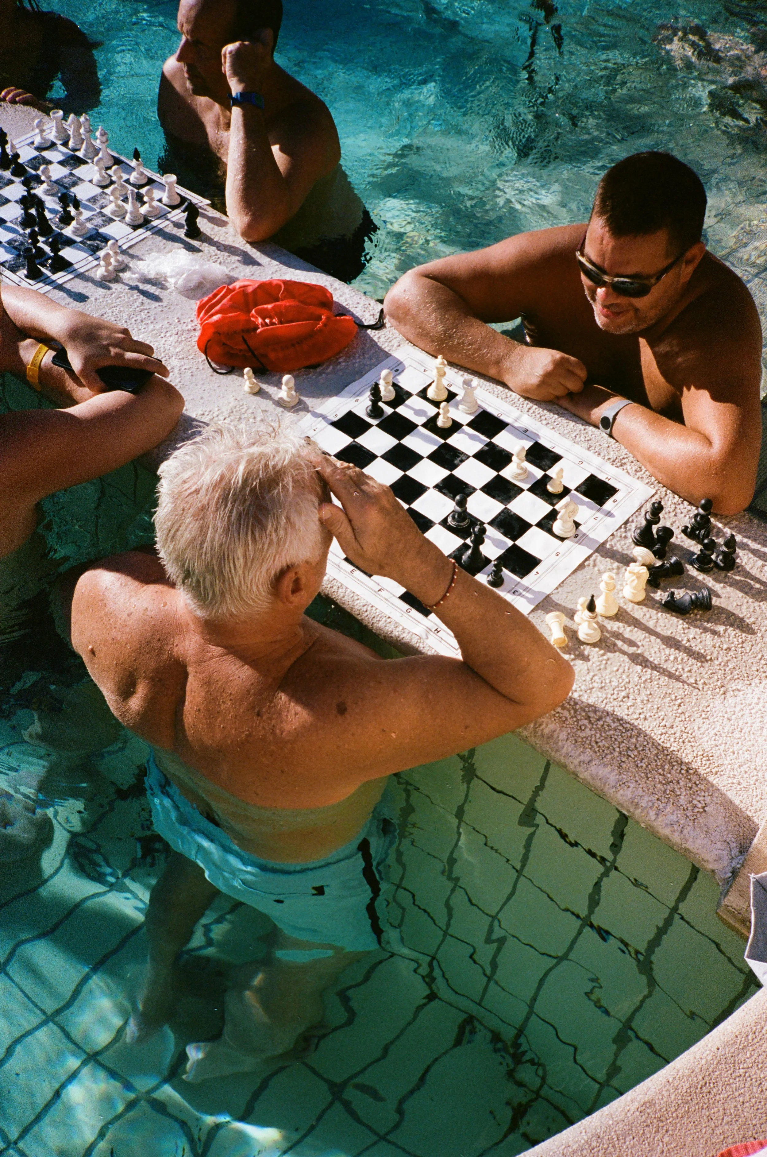 People playing chess on a table at the edge of a swimming pool.