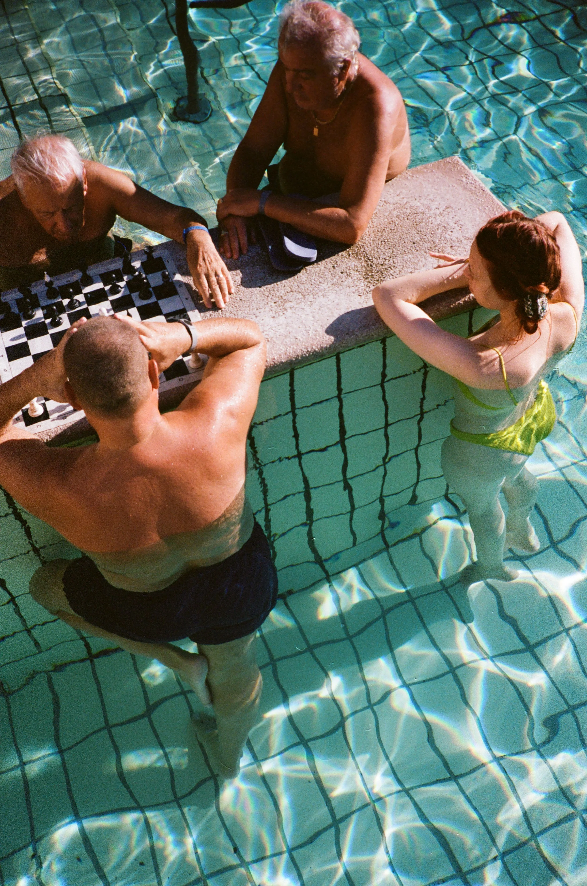 Two elderly men and one young woman are playing chess on a table at the edge of a swimming pool, with the woman and one of the men in the water.