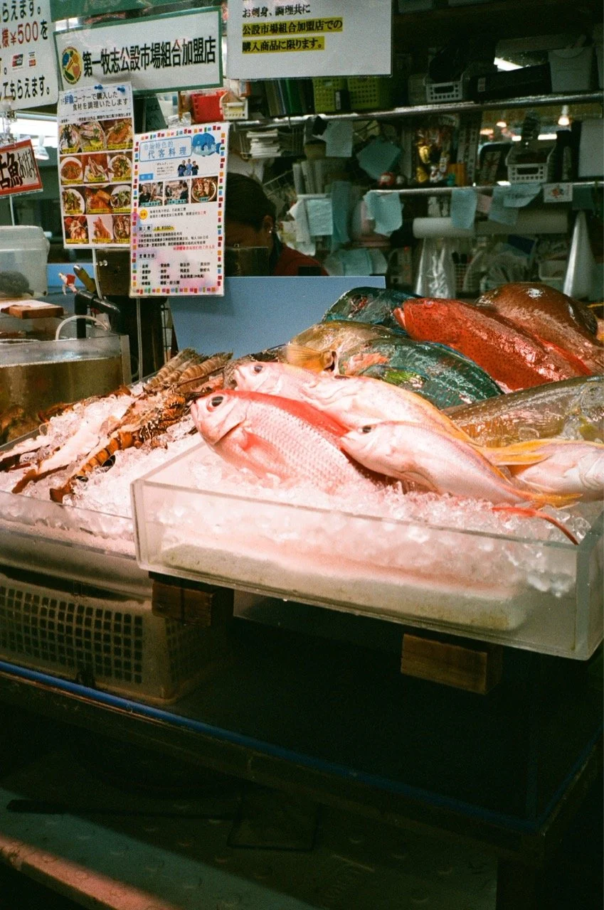 Fresh fish and seafood displayed on ice at a market stall with Japanese signs and menus in the background.