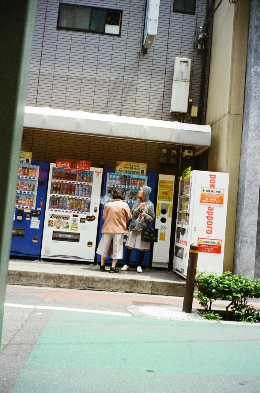 Two women stand near a row of vending machines outside a building, with a small green bush and a brown post nearby.