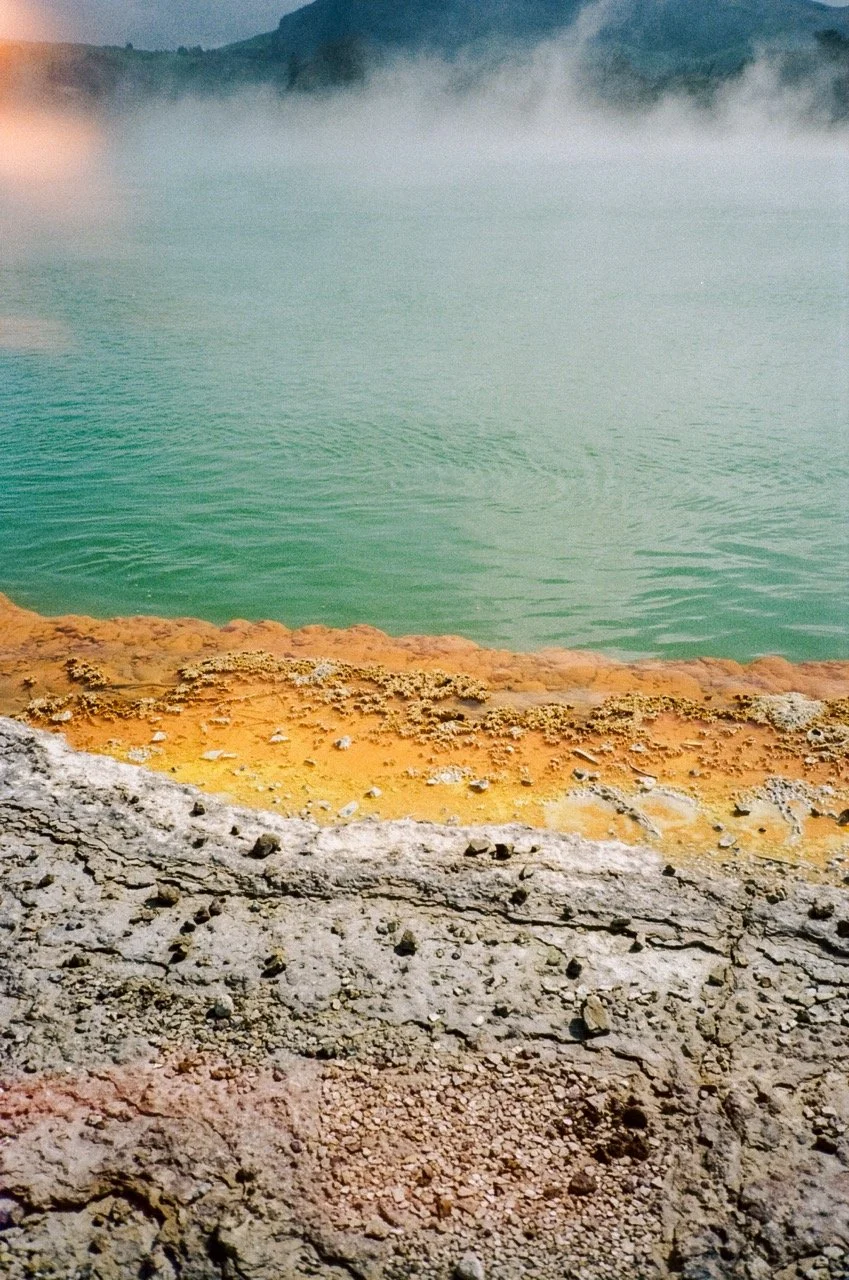 Colorful geothermal hot spring with mineral deposits and bubbling water, surrounded by steam and distant mountains.