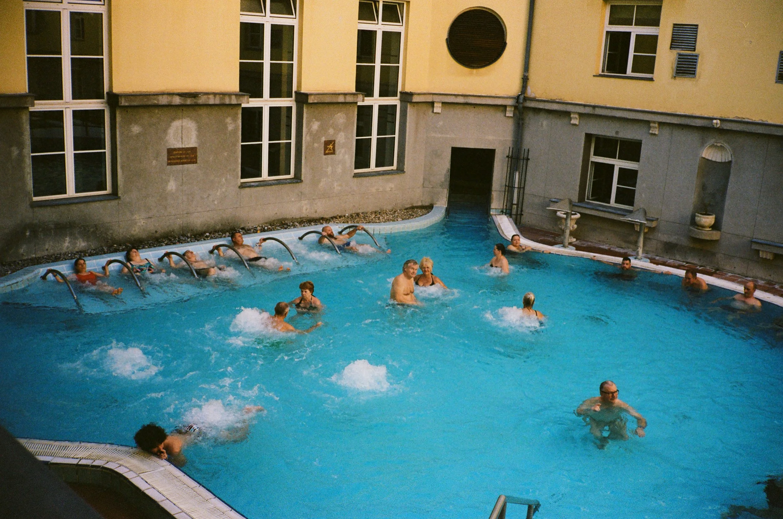 People swimming and relaxing in an indoor pool with a yellow exterior wall, multiple windows, and decorative water spouts.