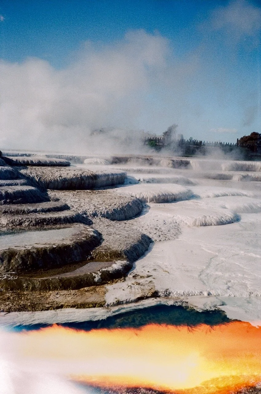 Steamed geothermal pools with white mineral deposits, blue sky, and a fence in the background.