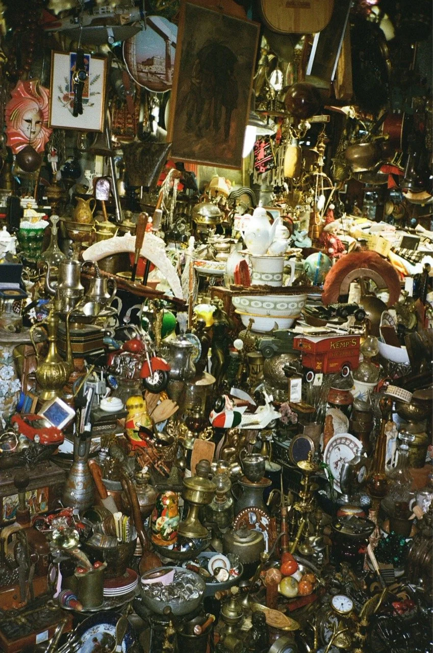 A dense collection of vintage and antique items including clocks, teapots, figurines, lamps, picture frames, and decorative objects in a cluttered space.
